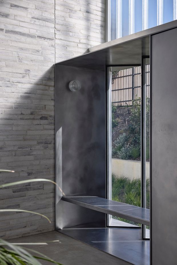 A narrow, light-filled modern interior corner with whitewashed exposed brick walls, dark polished concrete or metal surfaces, and a slim black-framed glazed door or window opening to a landscaped courtyard. A dark perforated built-in bench or shelf projects beneath the glazing, while strong sunlight creates geometric shadows across the walls and floor. The space has a minimalist, industrial character with tall glazing and restrained detailing.
