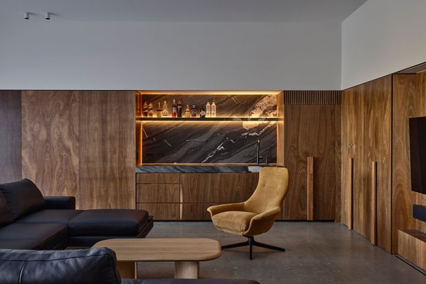 A sophisticated contemporary living room with extensive dark timber wall panelling and built-in cabinetry. A black leather sectional sofa faces a low rounded timber coffee table, while a sculptural tan lounge chair sits near a backlit built-in bar or display niche. The niche features bottle storage, warm LED lighting, and a dramatic dark veined stone backsplash. The room has a smooth grey concrete-look floor, recessed track lighting, and a wall-mounted television integrated into the timber joinery. The overall design is dark, refined, and luxurious.