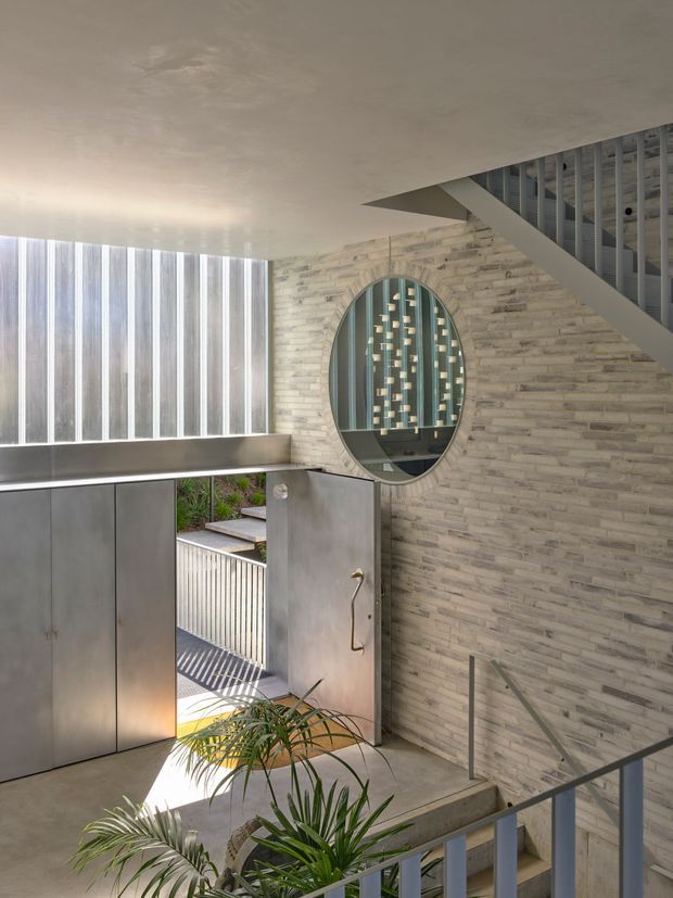 A contemporary interior stair hall or entryway with a split-level stair descending toward an open metal door. The space features a pale stacked-stone wall, a large oval internal window with decorative vertical elements, exposed stair framing, a smooth concrete ceiling and floor, and a slim metal handrail. Vertical translucent glazing brings in strong daylight, while a small palm adds greenery near the entrance.