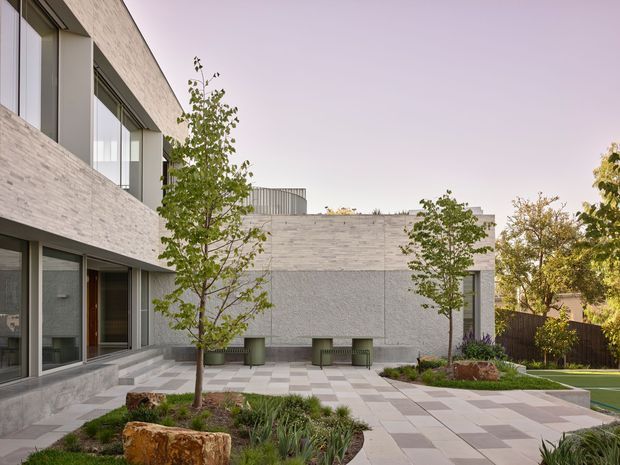 A contemporary residential courtyard viewed from outside, featuring pale brick and textured rendered walls, large dark-framed windows and sliding doors, a broad paved terrace with alternating light-grey tiles, landscaped garden beds with young trees and ornamental grasses, and a glimpse of a swimming pool at right. The low, rectilinear building forms and enclosed outdoor space create a calm, refined modern setting.