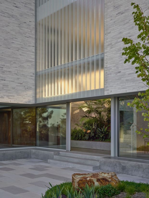 Exterior architectural view of a modern two-storey building with pale stacked-stone cladding, extensive floor-to-ceiling glazing, and a tall illuminated glazed opening screened by vertical fins. A recessed planted courtyard or indoor garden is visible behind the glazing, with concrete steps leading to a paved terrace and landscaped planting in the foreground.