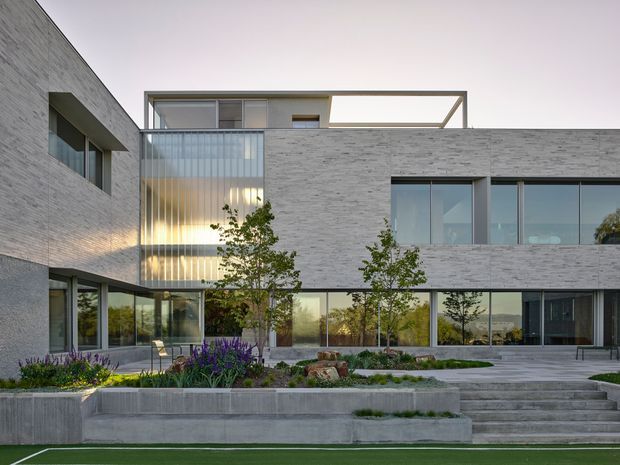 A contemporary multi-storey residence with a strong cubic composition, pale grey masonry cladding, extensive floor-to-ceiling glazing, and a recessed upper-level terrace or roof opening. The building forms a landscaped courtyard frontage with concrete retaining planters, broad steps, ornamental planting, and a level lawn. Warm interior lighting is visible through the glazed central section, while the architecture remains clean, restrained, and highly geometric.
