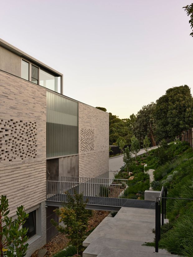 Contemporary multi-level residential building viewed from an elevated landscaped walkway. The facade combines pale horizontal brickwork, patterned perforated brick screens, dark metal framing and a tall translucent ribbed glazing panel. A pedestrian bridge and concrete path connect the split-level structure, with dense planting surrounding the site.