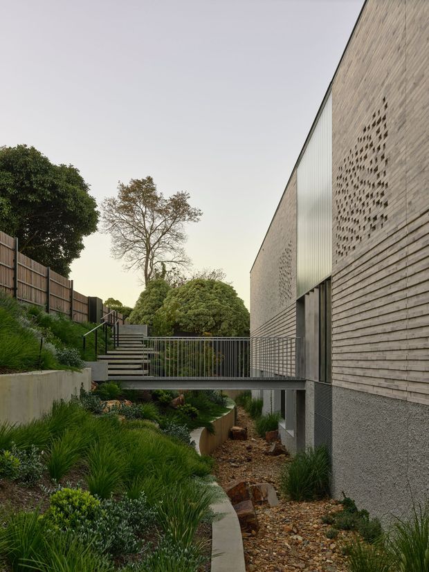 Exterior architectural view of a contemporary building set into a landscaped sloping site. A narrow pedestrian bridge and stairs connect through dense garden planting beside a dry creek bed. The facade combines pale textured masonry, patterned breeze-block openings, horizontal timber-like cladding, dark framed glazing, and a translucent vertical panel. Metal balustrades and restrained concrete retaining walls contribute to the minimalist, carefully integrated landscape design.