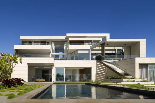 Exterior view of a large multi-level contemporary residence arranged around a rectangular swimming pool. The building features crisp horizontal and cantilevered volumes, pale stone or tiled cladding, extensive glazing, glass balustrades, recessed balconies, and a prominent external stair with dark metal framing and slatted screening. Landscaped planting and lawn surround the pool under a clear blue sky.