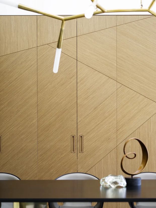 A refined dining area dominated by full-height light timber veneer cabinetry with geometric panel seams and integrated vertical handles. A dark dining table and curved chairs sit beneath a sculptural brass-and-white pendant light, with a small abstract decorative object on the table. The composition is warm, clean-lined, and contemporary with subtle mid-century influence.