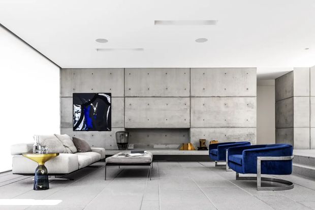 A bright, minimalist contemporary living room with a concrete feature wall, integrated low cabinetry, pale tiled flooring, a large white sectional sofa, and two vivid cobalt-blue lounge chairs with metal frames. A low glass coffee table and sculptural side table complete the refined, gallery-like space.