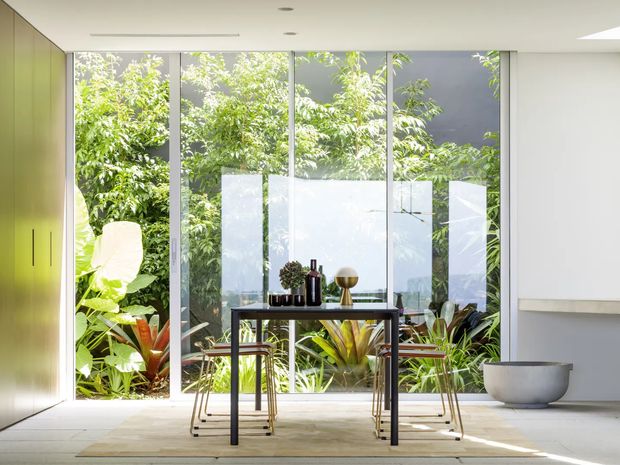 Bright minimalist dining area with a slim dark dining table and stools positioned in front of full-height glazed doors or windows. Lush planting is visible immediately outside, while pale flooring, white walls, integrated cabinetry, and a simple sculptural bowl create a clean indoor-outdoor atmosphere.