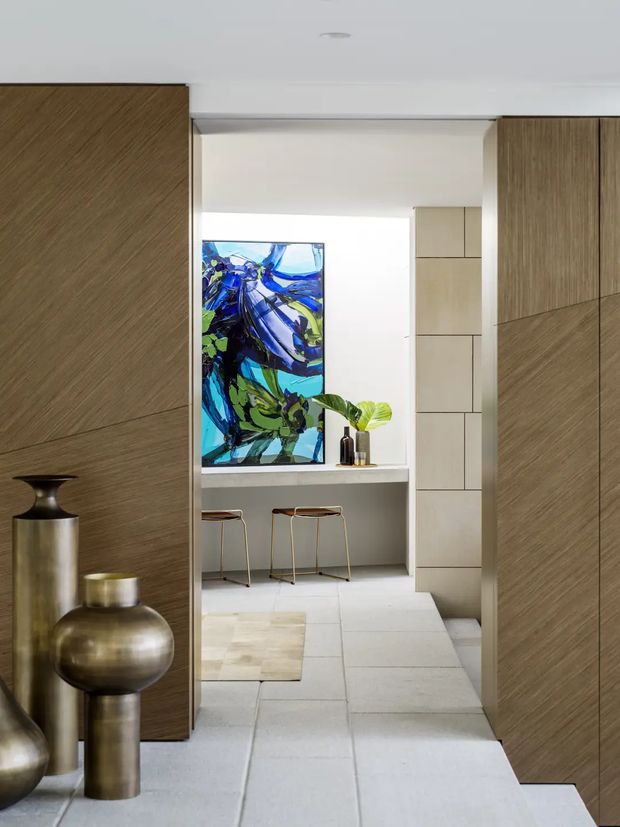 View through a wide opening framed by dark timber wall panelling into a bright contemporary kitchen or bar nook. The inner space has pale stone or tile flooring, a light benchtop with two slim stools, pale built-in cabinetry, a colourful abstract artwork, and decorative vessels with greenery. Large timber-panelled doors or walls dominate both sides of the foreground.