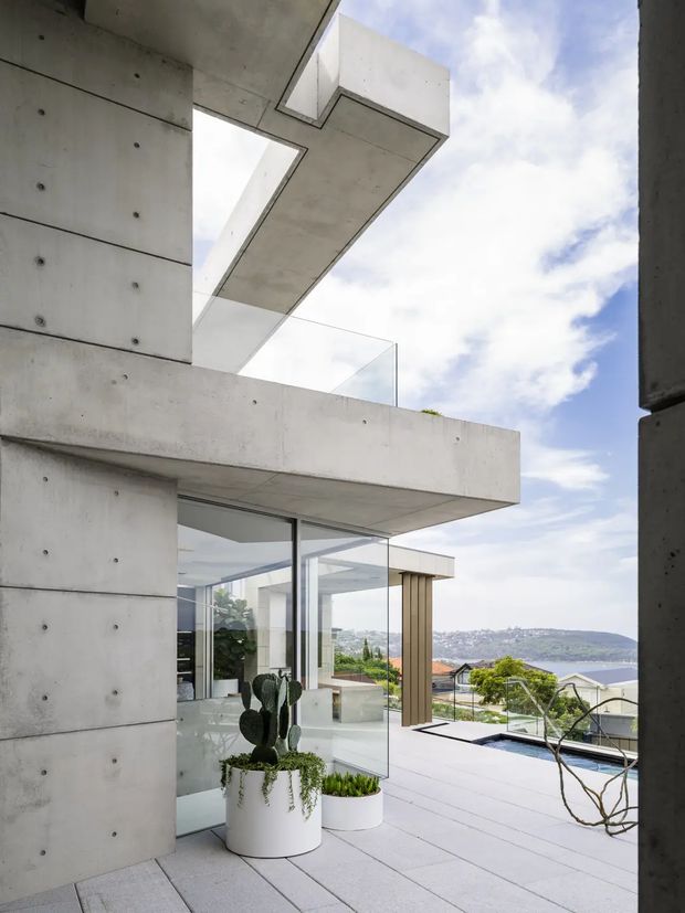 A striking view of a contemporary concrete residence with projecting cantilevered slabs, smooth board-marked concrete walls, extensive frameless glazing, and a broad tiled terrace. The terrace overlooks a bay and hilly urban landscape, with potted cacti and sculptural planting near the glass-walled interior. The composition emphasises strong geometric forms, open sky, and indoor-outdoor connection.