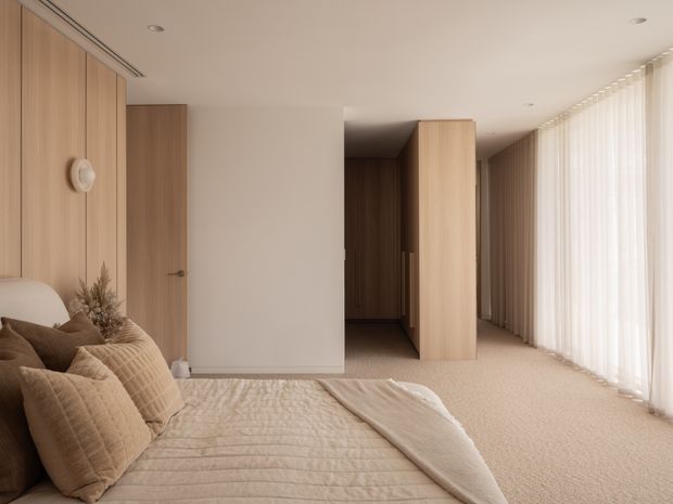 A warm, minimalist bedroom with a low bed in the foreground, layered beige quilted bedding and cushions, and full-height sheer curtains along the right side. Light timber wall panelling and matching built-in doors create a continuous joinery treatment, with a softly lit dressing or wardrobe passage beyond the central opening. The room has recessed ceiling lights, a neutral palette, and a calm, refined atmosphere.