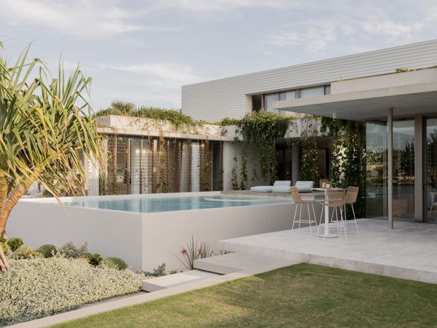 A contemporary flat-roofed residence viewed from its landscaped outdoor area, with clean horizontal lines, pale cladding, extensive glazing, a raised rectangular swimming pool, pale paved terraces, and lush subtropical planting. The image emphasises the modern exterior architecture and pool terrace.