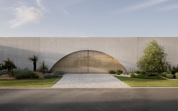 Front exterior view of a minimalist contemporary building with a long, smooth pale concrete facade and a distinctive large arched entrance enclosed by vertical metal screening. A light paved driveway leads from the street to the entrance, with manicured lawns and drought-tolerant landscaping on both sides.