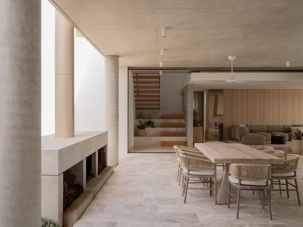 A refined open-plan living and dining area with a minimalist concrete fireplace, large stone-look flooring, a sculptural dining table with woven chairs, and a warm timber-clad kitchen or living joinery wall. A partially enclosed stairway with timber slats and integrated planting is visible in the background. The space has a calm, neutral palette and strong indoor-outdoor openness.