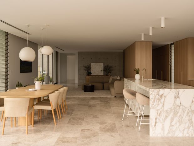 Open-plan contemporary interior combining a dining area, kitchen and living room. The space features large-format beige stone-look flooring, warm timber cabinetry, a substantial veined marble or stone kitchen island, upholstered dining chairs and bar stools, pendant globe lighting, and a neutral-toned lounge area. Full-height glazing and louvred window elements bring in natural light, while the restrained palette creates a refined, warm atmosphere.