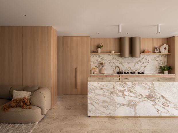 Warm, refined open-plan kitchen with full-height timber cabinetry, a large veined stone island, matching stone splashback, integrated cooktop and sink, and sculptural cylindrical rangehoods. A curved upholstered lounge chair with a small dog sits to the left, and subtle shelving decor adds softness.
