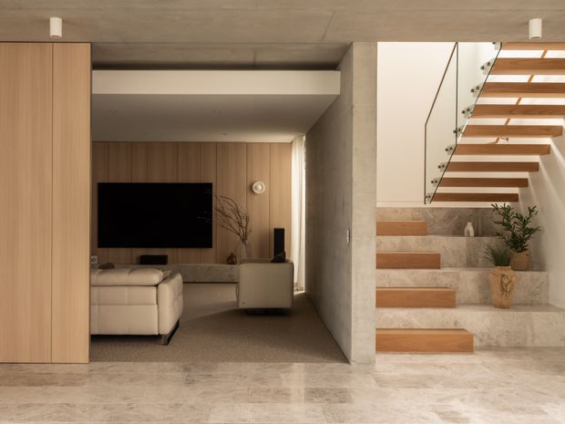 Interior view of a refined open-plan living room and stairway. The living area features full-height light timber cabinetry and wall panelling, a wall-mounted television, a pale upholstered sofa, and a low built-in console. Beside it, a floating timber staircase with a slim glass balustrade rises alongside a textured pale stone or concrete wall. The space has polished pale stone flooring, warm neutral tones, recessed lighting, and a minimalist contemporary character.