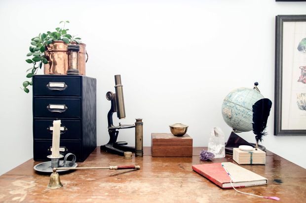 A styled vintage desktop vignette featuring a worn timber desk, dark filing drawers, copper planter, antique microscope, globe, framed artwork, stacked books, candleholder, and assorted small objects against a white wall.