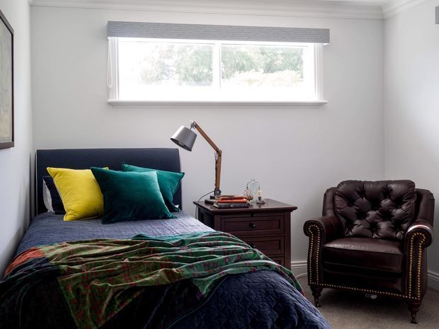 A compact, traditionally styled bedroom with a single bed beneath a wide window, layered in dark blue bedding and a green patterned throw with bright yellow and teal cushions. A dark timber bedside table supports an angled desk lamp, while a deep brown tufted leather armchair sits beside it. White walls, classic cornice detailing, a grey roller blind, and neutral carpet complete the room.