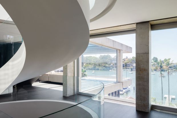 Contemporary waterfront interior featuring a dramatic sculptural curved white staircase or architectural feature, polished dark flooring, exposed concrete columns, glass balustrades, and extensive floor-to-ceiling glazing overlooking a marina and lush shoreline. The space is bright, open, and highly sculptural, with a refined modern aesthetic.