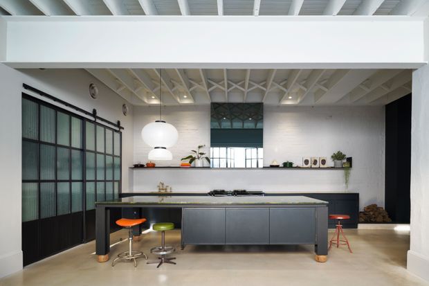 A bright, open-plan contemporary kitchen with a large central island, dark grey built-in cabinetry, integrated cooktop, and several stools. The space features white painted brick walls, exposed white timber roof framing and beams, polished concrete flooring, industrial-style black-framed glazed sliding doors, open shelving, and a large sculptural pendant light.