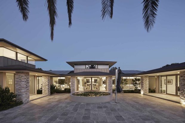 A low-rise contemporary residential courtyard viewed from outdoors at dusk. The U-shaped building has broad glazed openings, dark low-pitched roof planes, pale stacked-stone wall sections, and a central circular raised planter or water feature. Large-format grey paving extends through the courtyard, with palm trees framing the scene and warm interior lighting visible through the glazing.