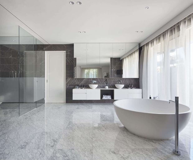 Bright, spacious contemporary bathroom with a freestanding white oval bathtub, twin floating vanities, large mirrored cabinetry, dark marble-look wall tiles, and pale polished stone flooring. Full-height sheer curtains and extensive glazing bring in abundant natural light, while a frameless glass shower enclosure occupies the left side.