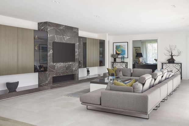 Bright contemporary living room with a large grey sectional sofa arranged around a low coffee table and a marble-effect feature wall incorporating a television and linear fireplace. Full-height timber-toned built-in cabinetry, pale flooring, recessed lighting, artwork, and a small adjoining desk area complete the refined open-plan interior.
