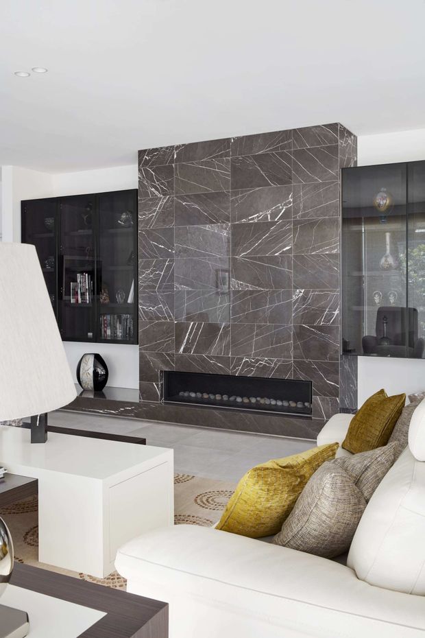 A contemporary living room centred on a dramatic dark grey, white-veined stone feature wall with a long built-in fireplace. Black glazed display cabinetry flanks the feature wall, while a white sectional sofa with mustard-gold and grey cushions faces a white coffee table. The room has a light neutral floor, recessed ceiling lights, and a monochromatic palette with warm yellow accents.