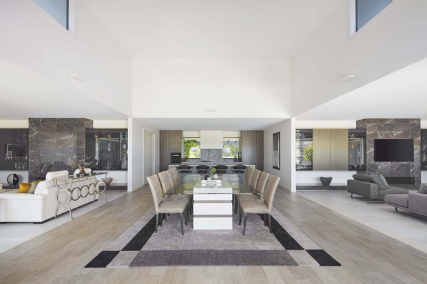 Bright, spacious open-plan contemporary interior combining a dining area, kitchen and living room. The room features a long dining table with upholstered chairs, pale timber-look flooring, white walls and cabinetry, dramatic dark marble-look feature walls, recessed lighting, and large windows. A lounge area with a television and sofas is visible on the right, while additional seating and joinery appear on the left.