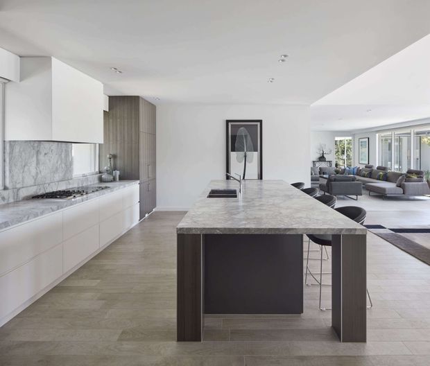Contemporary open-plan kitchen with a large marble-look island, white flat-panel cabinetry, a grey veined stone splashback and integrated cooktop. Dark timber-toned island supports and cabinetry contrast with the pale flooring and white walls. A furnished living area with grey sofas is visible beyond the kitchen.