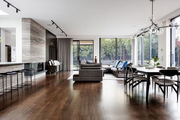 Bright, open-plan contemporary living space combining a kitchen, dining area and lounge. The room features dark timber flooring, a large stone-clad fireplace wall with integrated television, extensive floor-to-ceiling glazing, a grey sectional sofa, black dining furniture, and modern track and pendant lighting. The space has a refined, spacious feel with views to greenery outside.