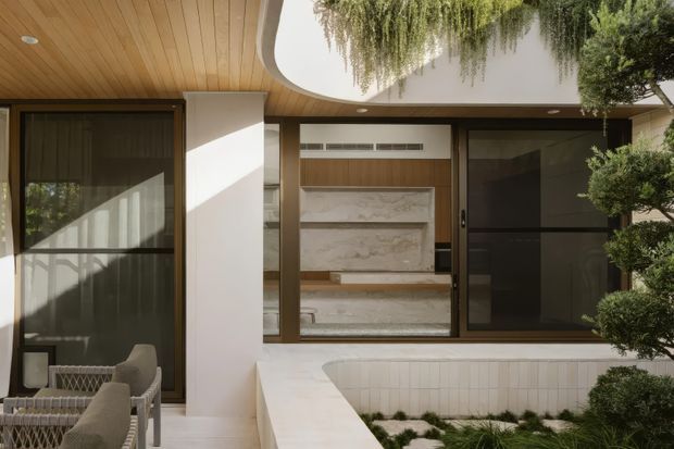A refined courtyard-facing exterior/interior threshold with dark-framed glazed openings, warm timber soffit, curved white plaster ceiling detail, and a marble-clad interior feature wall visible through the glazing. A pale tiled raised planter or ledge separates the space from clipped ornamental greenery, while outdoor seating occupies the lower left.