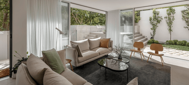 A bright, contemporary living room with floor-to-ceiling glazing opening onto a landscaped courtyard. The room contains a large neutral sectional sofa, a second beige sofa, sculptural timber lounge chairs, a black oval glass coffee table, and a dark area rug. White sheer curtains, pale walls, and expansive black-framed windows create a calm indoor-outdoor connection, while concrete steps, pale paving, and planted walls are visible outside.