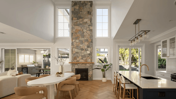 A bright, open-plan contemporary interior combining a kitchen, dining area and living room. The space features a double-height ceiling, tall multi-pane windows, a central rough natural-stone fireplace chimney, white walls and cabinetry, warm herringbone timber flooring, and a large white stone kitchen island with timber bar stools. Neutral upholstered seating and dining furniture create a refined, airy atmosphere, with abundant greenery visible through the glazing.