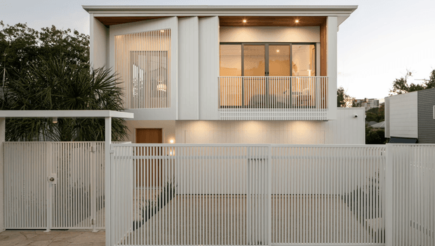 Front exterior view of a modern two-storey residence with a crisp white facade, deep upper-level balcony, vertical metal screening, large glazed openings, and a secure slatted driveway gate. Warm concealed and facade lighting highlights the entry and balcony at dusk. Tropical planting frames the left side, while neighbouring urban buildings are visible in the background.