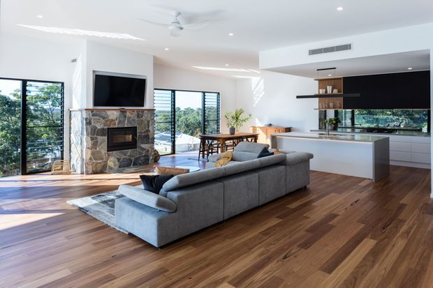 Bright open-plan living area with a large grey sectional sofa, stone-clad fireplace and wall-mounted television. The space includes timber flooring, expansive black-framed glazing, a dining area, and a contemporary kitchen with a central island and dark built-in cabinetry.