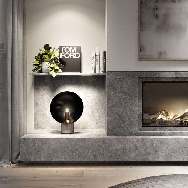 A refined contemporary living room feature wall with a built-in fireplace surround and recessed display niche. The wall is clad in mottled grey stone or stone-look finish, with a black inset firebox, sculptural black table lamp, plant, books, artwork and soft grey curtains. Pale timber flooring and a partial rug add warmth to the otherwise monochromatic scheme.