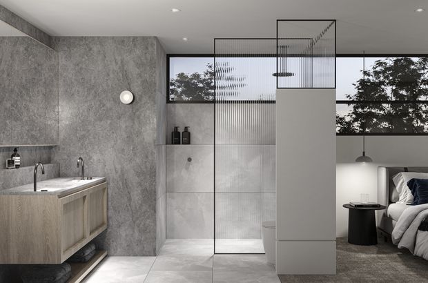 A sophisticated open-plan ensuite bathroom and bedroom interior. The bathroom features a floating light timber vanity with integrated basin, black tapware, large grey stone-look wall and floor finishes, and a frameless-style ribbed glass shower enclosure. A partial-height dividing wall separates the bathroom from a contemporary bedroom with a dark upholstered bed, pendant lighting, and large horizontal windows. The overall scheme is monochromatic, refined, and minimalist with black metal accents.