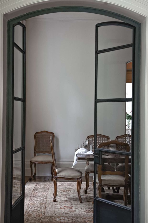 View through tall black-framed glazed double doors into an elegant dining room. The room has pale walls with decorative moulded trim, a patterned Persian-style rug, a small dining table set with glassware and white linens, and several traditional cane-back wooden chairs. The arched doorway and dark metal-framed glazing create a strong architectural focal point.