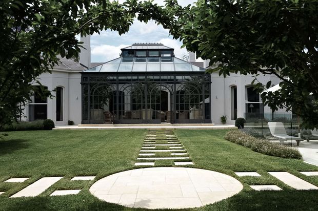 Formal garden approach to a white, symmetrical residence with a prominent glazed conservatory or glasshouse pavilion. The dark metal-framed structure has arched glazing and a raised glazed roof, while a geometric stepping-stone path crosses the manicured lawn to a circular pale-paved landing. Mature trees frame the view, with restrained planting and outdoor seating visible at the sides.