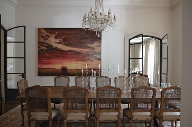 Elegant formal dining room with a long dark-wood dining table surrounded by woven-back upholstered chairs, crystal candelabra and a large ornate chandelier. White walls with detailed classical cornices and panelled trim frame black steel or dark-framed glazed arched doors, while a large atmospheric landscape artwork provides a deep red and brown accent.