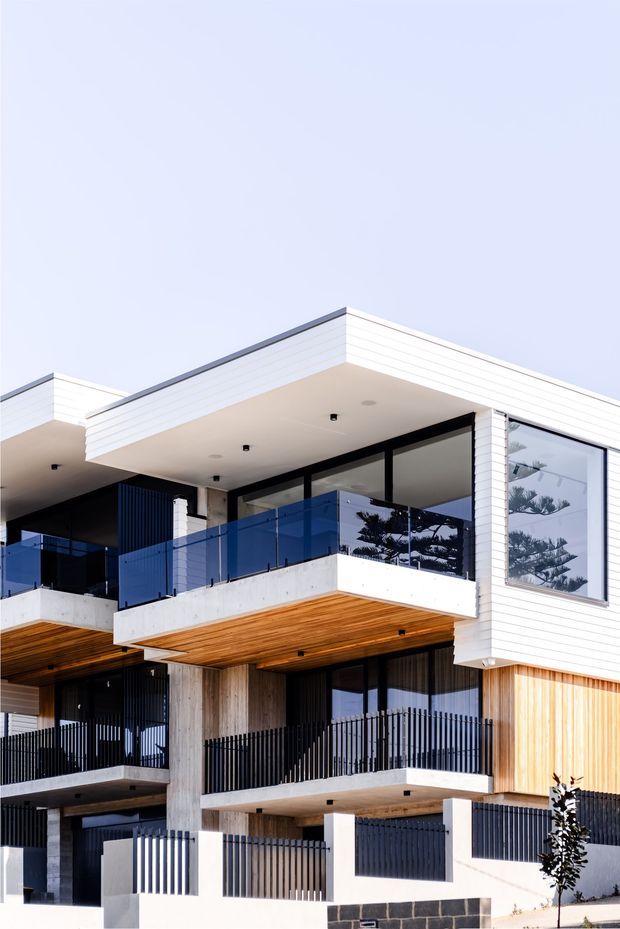 Contemporary multi-level residential building exterior with crisp white horizontal cladding, projecting flat roof slabs, deep balconies, dark metal balustrades, expansive black-framed glazing, exposed concrete columns, and warm timber soffits and wall accents. The facade has a clean geometric composition with blue-tinted glass balcony panels and a bright, refined coastal-residential appearance.