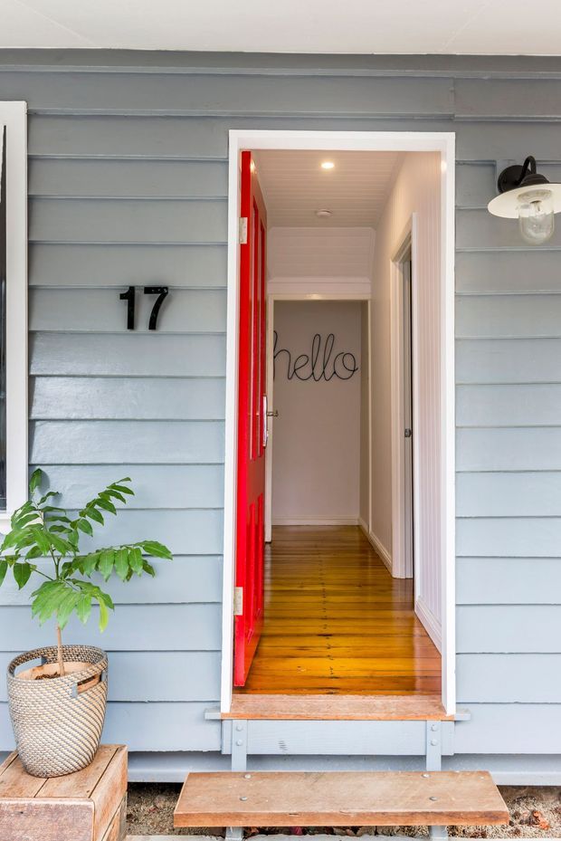 Front entry of a small timber-clad home, featuring pale blue-grey horizontal weatherboards, a vivid red open door, white trim, warm polished timber flooring, and a simple covered porch with a potted plant and wall-mounted exterior light. The hallway beyond has white panelling and a handwritten-style “hello” wall detail.