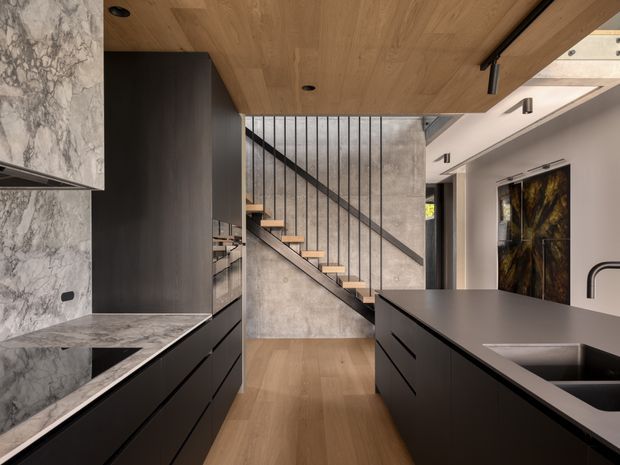 A contemporary open-plan kitchen viewed toward a stairway. The room features dark matte cabinetry, a large dark island with integrated sink, grey marble-look stone benchtops and splashback, warm timber flooring and a timber-lined ceiling. A concrete-textured wall and slim black metal stair balustrade create an industrial architectural feature, with floating timber stair treads leading upward.