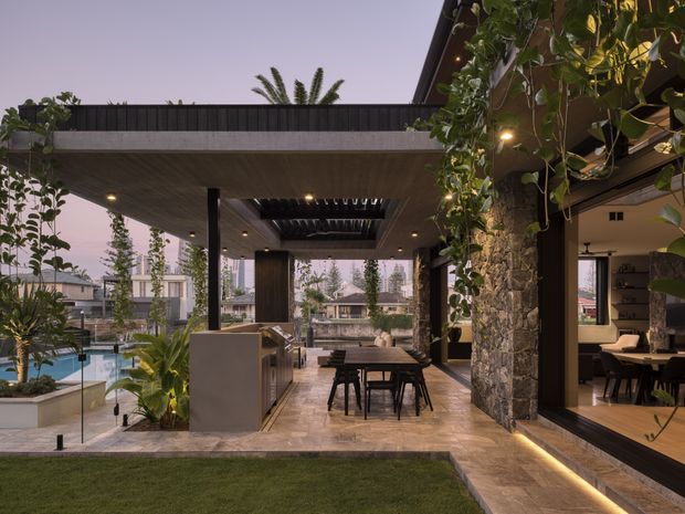 A sophisticated outdoor entertaining area adjoining a contemporary home, with a covered patio, dining table, outdoor kitchen or bar, swimming pool, glass balustrade, and lush integrated planting. The space features a flat-roofed timber-lined canopy, stone-clad structural columns, large openings, warm recessed lighting, and views into an adjoining interior dining or living space. The setting appears suburban and the atmosphere is warm and refined at dusk.