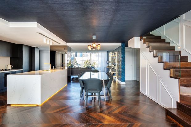 Open-plan contemporary interior combining a kitchen, dining area and living space. The room features dark herringbone timber flooring, a large white stone island with integrated warm underlighting, dark cabinetry, a deep charcoal textured ceiling, white wall panelling, and a timber staircase with glass balustrade. A blue patterned feature column and glazed doors add contrast.
