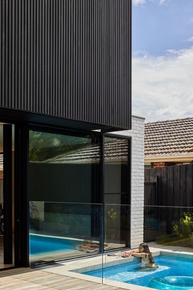 Exterior view of a contemporary dark-clad building beside a small bright-blue swimming pool. The projecting upper volume has closely spaced vertical black slats, while the ground floor uses extensive dark-framed glazing reflecting the pool and tiled roof of the neighbouring building. A white-painted brick wall, dark timber fence, pale paving, and pool glass balustrade complete the scene.