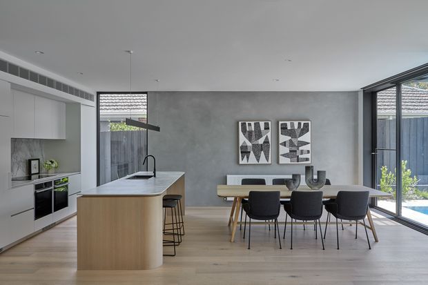 Open-plan contemporary kitchen and dining area with a pale timber island, white integrated cabinetry, grey concrete-look feature wall, light timber flooring, and a large dining table positioned beside full-height glazed doors. Minimalist monochrome artwork and neutral furnishings create a clean, refined interior with strong indoor-outdoor connection.
