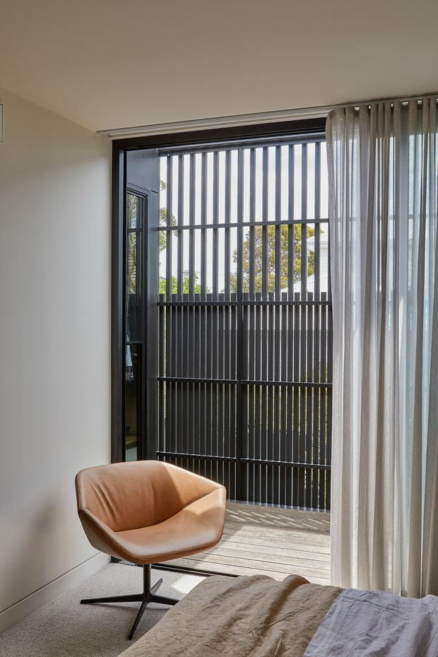 A calm, light-filled bedroom corner opening onto a small timber balcony or terrace. The room has neutral painted walls, carpeted flooring, sheer full-height curtains, a tan leather swivel armchair, and a partially visible bed with neutral bedding. A large glazed opening with dark framing leads outside, where vertical dark screening forms a privacy balustrade.